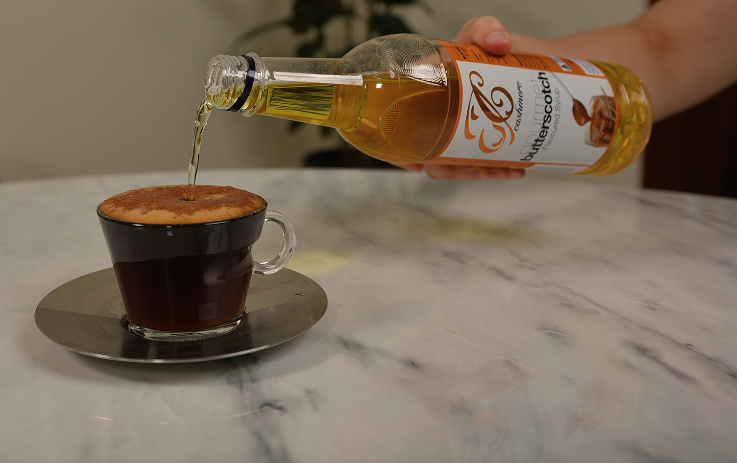 A person off screen drizzles a bottle of Cashmere Syrups Butterscotch Syrup into a cup of coffee on a white marble tabletop.